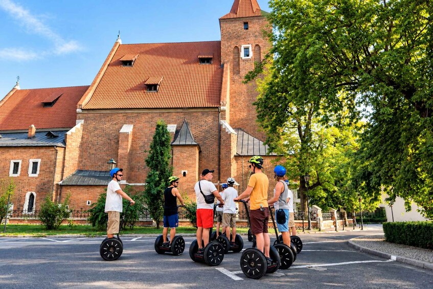Ryhmä ihmisiä ajaa segway-lautoilla historiallisesti merkittävän punatiilikoristeisen rakennuksen edessä, jonka katto on punainen. 5