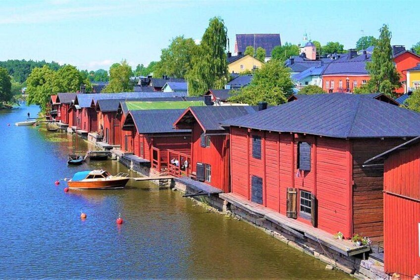 A row of red wooden buildings by a calm river, boats docked nearby, and a town in the background. 4