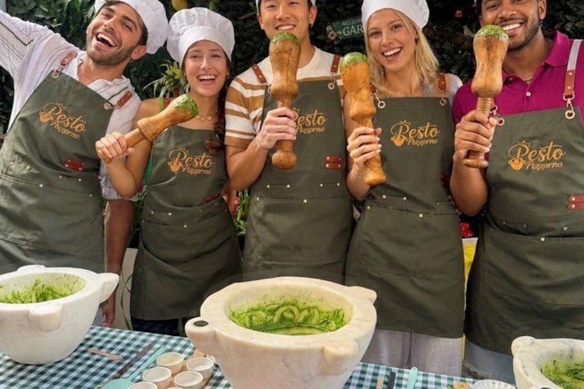 Eine Gruppe von Personen mit Schürzen, auf denen das Logo 'Pesto Pizzeria' zu sehen ist, hält Holzlöffel. Auf einer karierten Tischdecke stehen Schüsseln mit grünem Pesto. 1