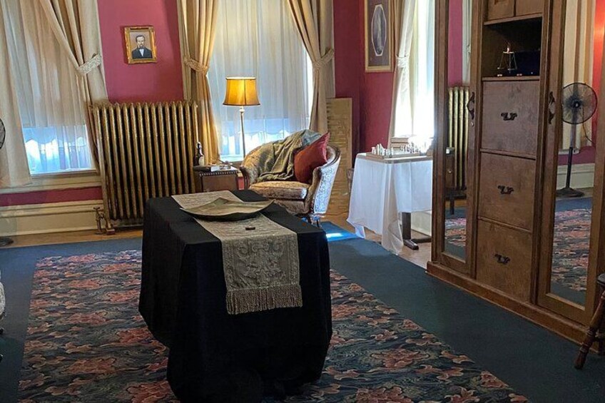 A room with a table covered by a tablecloth, a chair, a radiator, a lamp, and a cabinet. 2