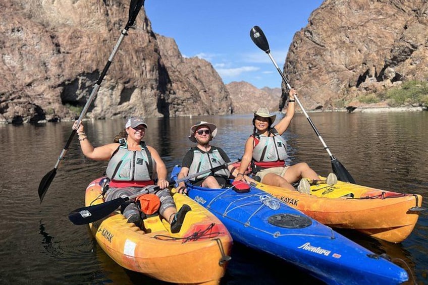 Tres personas remando en kayak por un río tranquilo con acantilados rocosos al fondo. 1