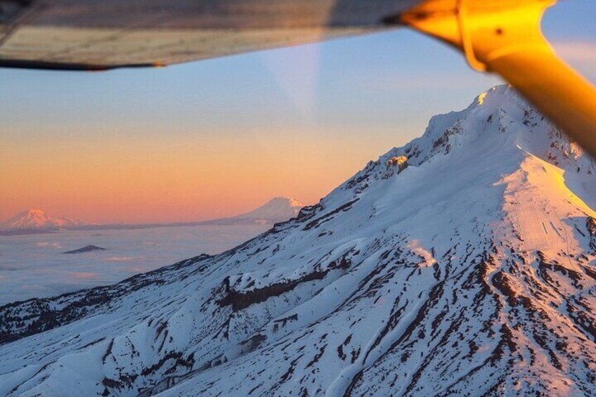 A snow-covered mountain at sunset with a plane's wing in the foreground. 1