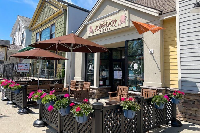 A building with a sign that reads 'AOMONDAK WINERY'. There are outdoor tables and chairs with umbrellas, and potted plants with pink flowers. 2