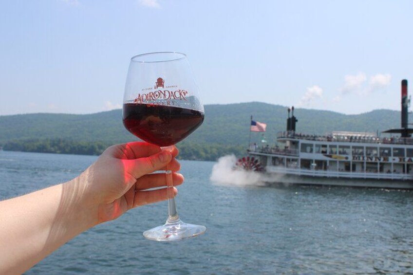 A hand holding a wine glass with a label, a boat with an American flag, and a body of water with hills in the background. 4