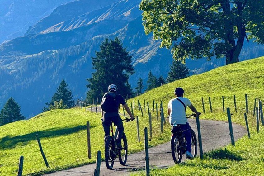 Two cyclists on a paved path with a mountainous landscape in the background. 1