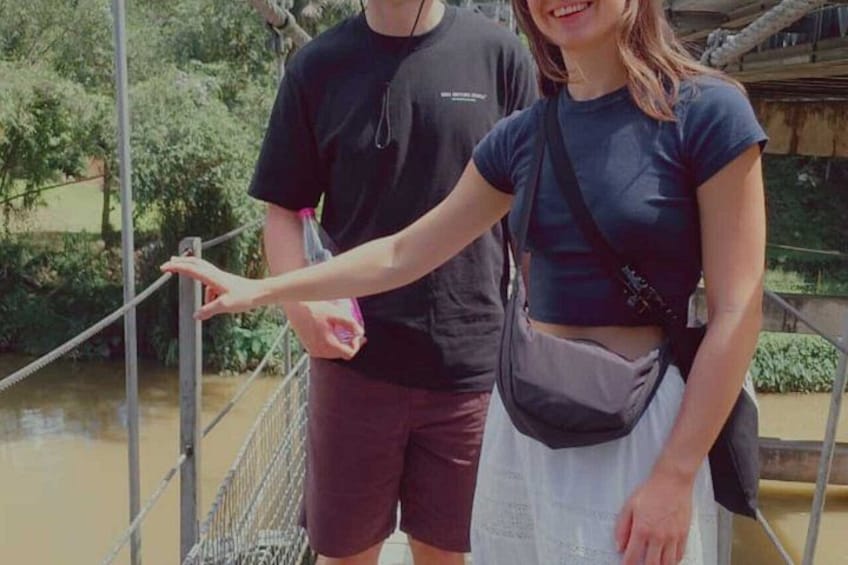 A person standing on a suspension bridge with a river below, wearing a dark top and carrying a bag. 1