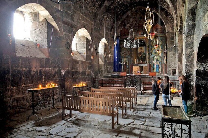 An ancient church interior with stone walls, wooden benches, and lit candles. 2