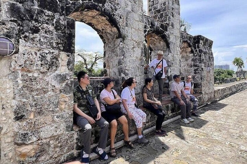 A group of people sitting on a stone wall with arches, a paved area, and a clear sky. 2
