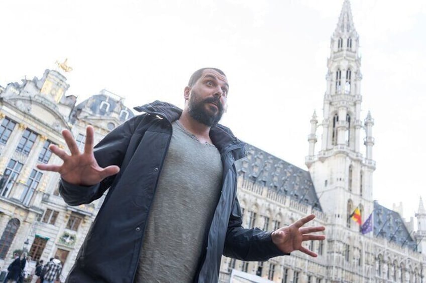 A person in a black jacket and gray shirt standing in front of a historic building with a tall tower. 1