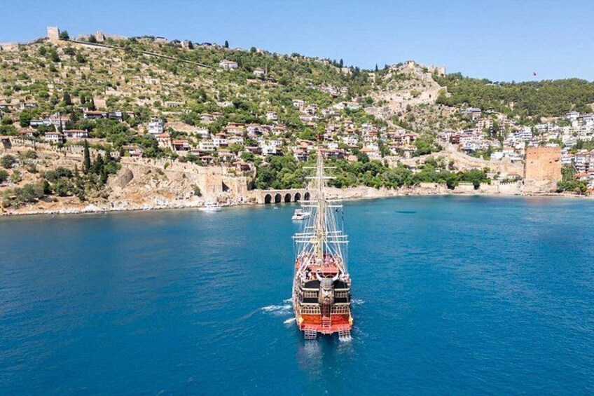 A sailing ship on a calm sea with a coastal town and a hill with a castle in the background. 4
