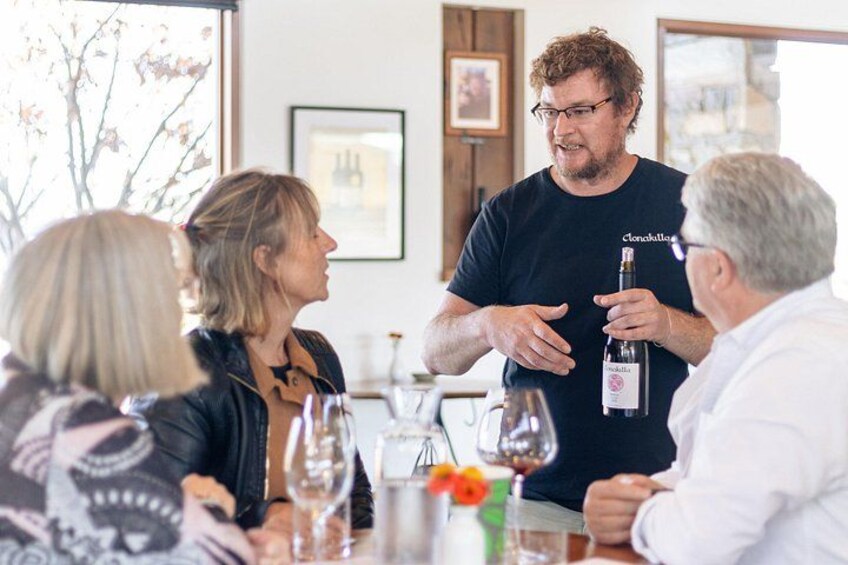 Un grupo de personas sentadas en una mesa con copas de vino, una botella y un jarrón con flores. 4