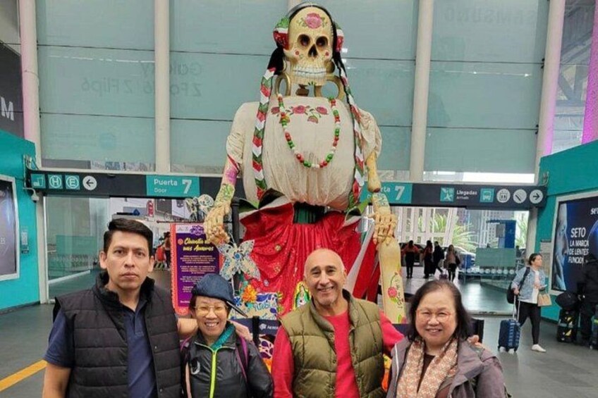 A large skeleton figure in a public indoor space, possibly a train station or airport, with a sign indicating 'Puerta 7' and 'Lingadas'. 4