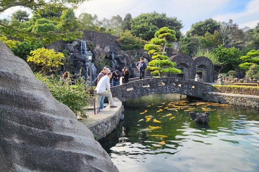 Un pont de pierre enjambe un étang où nagent des carpes koï, le tout étant entouré d’une végétation luxuriante et d’arbres. 3