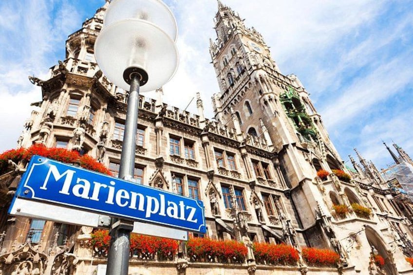 Ein Straßenschild mit der Aufschrift 'Marienplatz' vor einem historischen Gebäude mit aufwendiger Architektur. 1