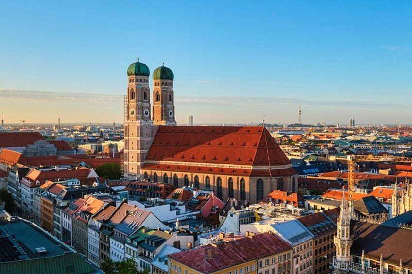Eine Stadtansicht mit einem markanten Kirchengebäude, das zwei grüne Kuppeln und einen Glockenturm zeigt, umgeben von historischen Gebäuden mit roten Ziegeldächern. 3
