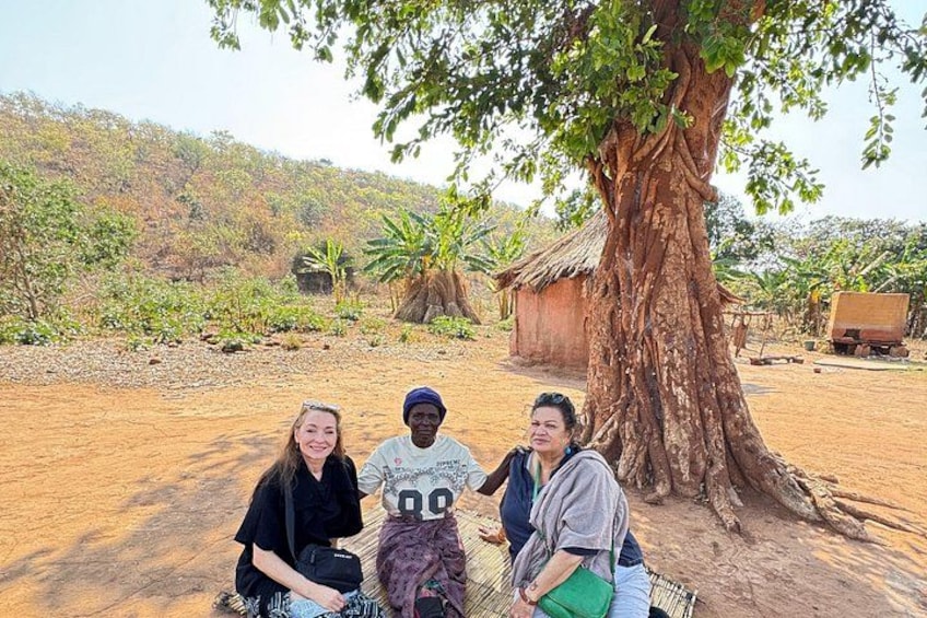 Three people sitting under a large tree in a rural setting with a dirt ground and a thatched-roof structure in the background. 2
