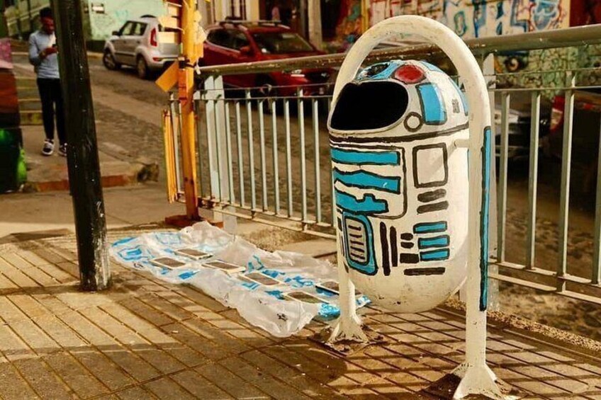 A sidewalk with a white and blue robot figure, a metal fence, and a plastic bag on the ground. 3