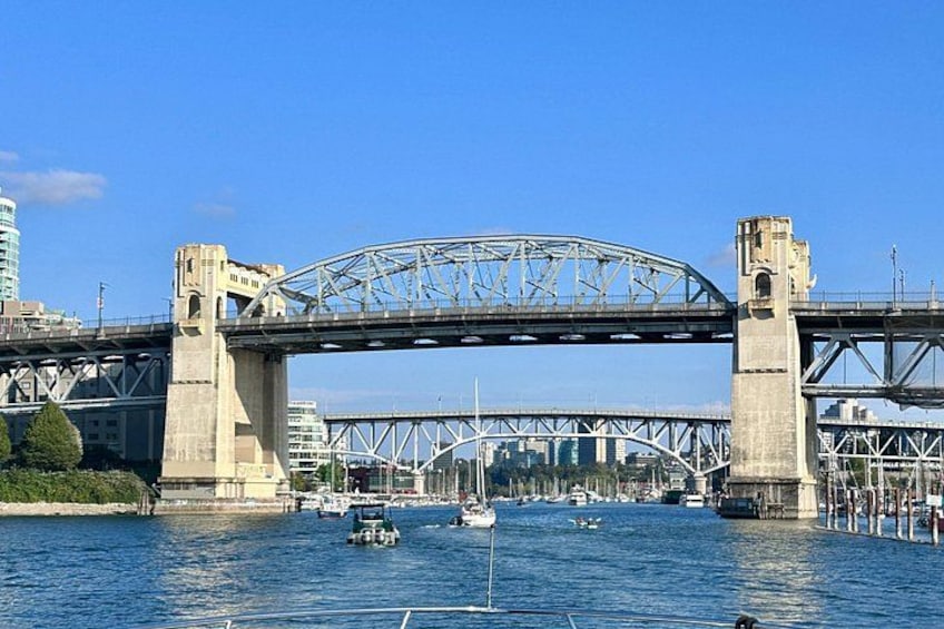 Een lange stalen brug over een wateroppervlak, met boten en stadsgebouwen op de achtergrond. 2