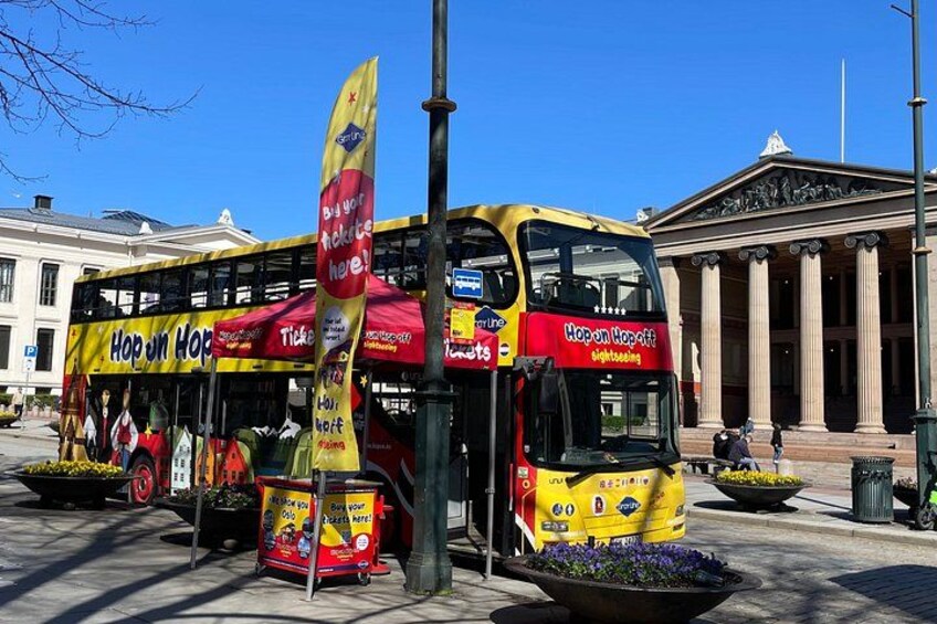 A double-decker bus with 'Hop on Hop Off' branding parked in front of a building with columns. 3