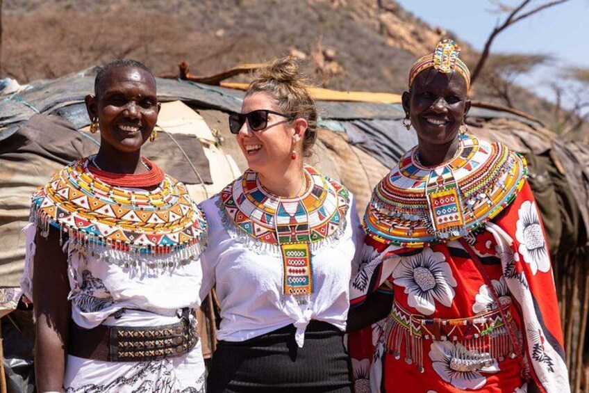 Three individuals in traditional attire with intricate beadwork and patterns, standing in front of a structure with a natural landscape in the background. 5