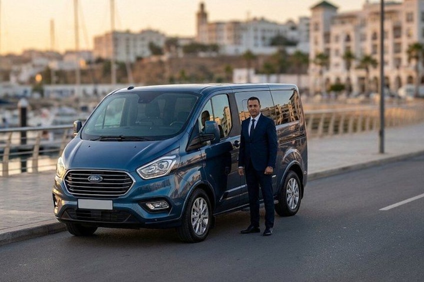 A blue van parked on a waterfront road with a cityscape in the background. 1