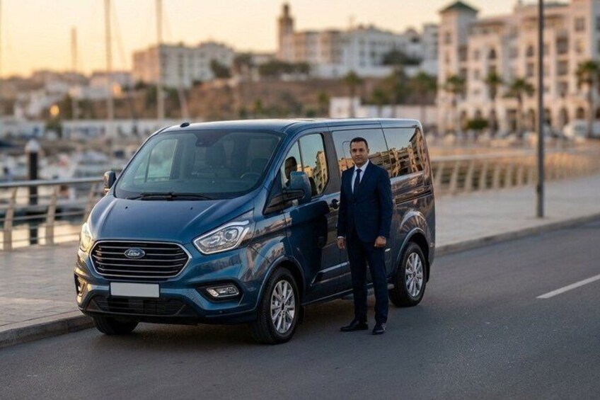 A blue van parked on a waterfront road with a cityscape in the background. 2