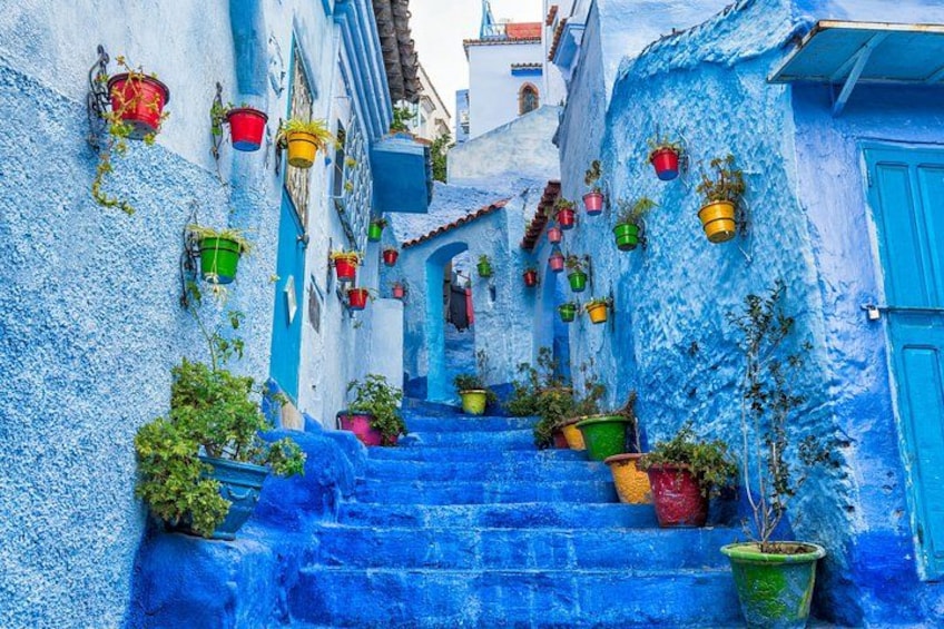 A narrow street with blue-painted buildings and colorful hanging planters. 2