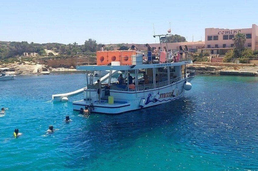 A boat with people swimming in the water, a clear blue sea, and buildings in the background. 2