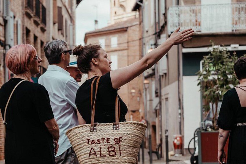 A person pointing towards a building, with a basket labeled 'TASTE OF ALBI' and a narrow street with buildings. 5