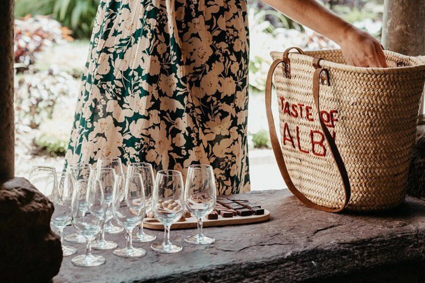A person in a floral dress holding a basket labeled 'Taste of ALB' with empty wine glasses on a stone surface. 2