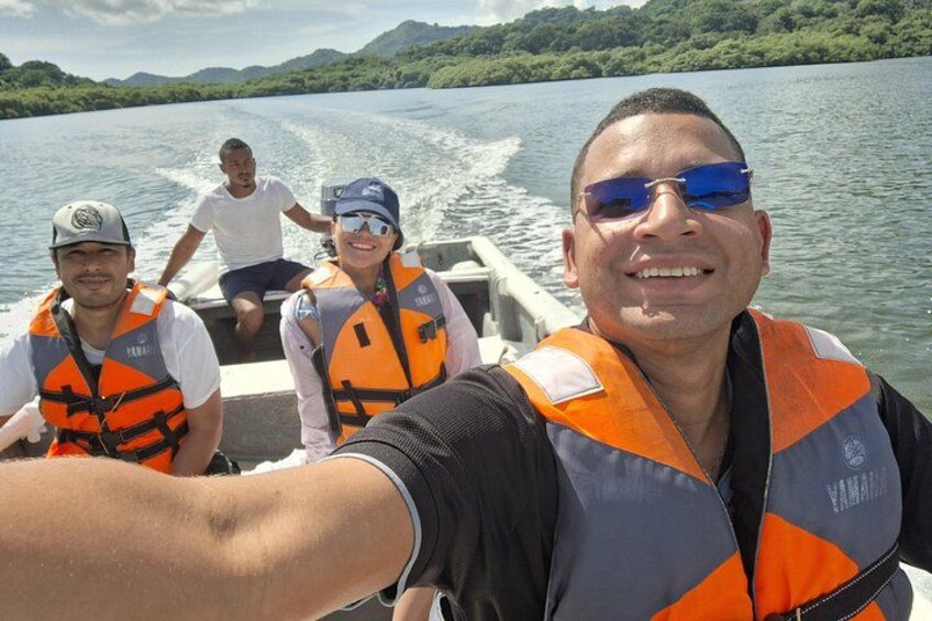 A group of people on a boat wearing life jackets, with a lake and greenery in the background. 5
