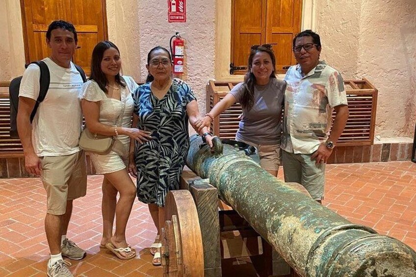 A group of people standing next to a large cannon in an outdoor setting with brick flooring and wooden doors in the background. 4