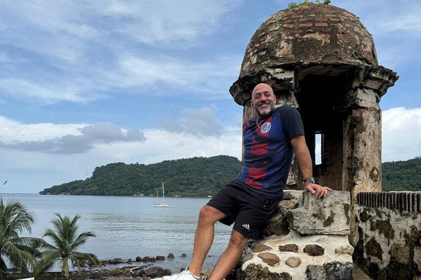 A man sitting on a stone structure by a body of water with a dome-shaped building, palm trees, and a distant island. 3