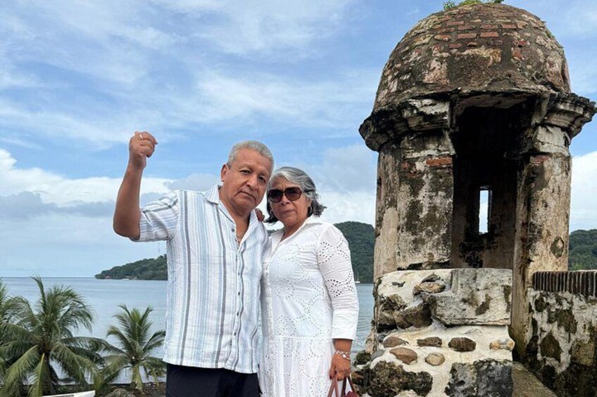 A man and woman standing in front of a historical stone structure with a clear view of the ocean and a distant island. 2