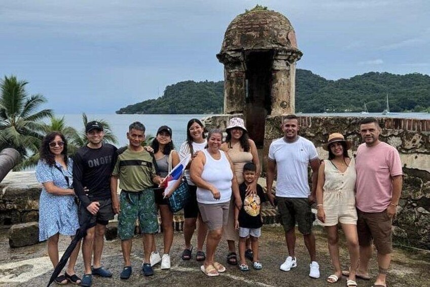 A group of people standing in front of a historical structure with a view of the sea and a hill in the background. 4