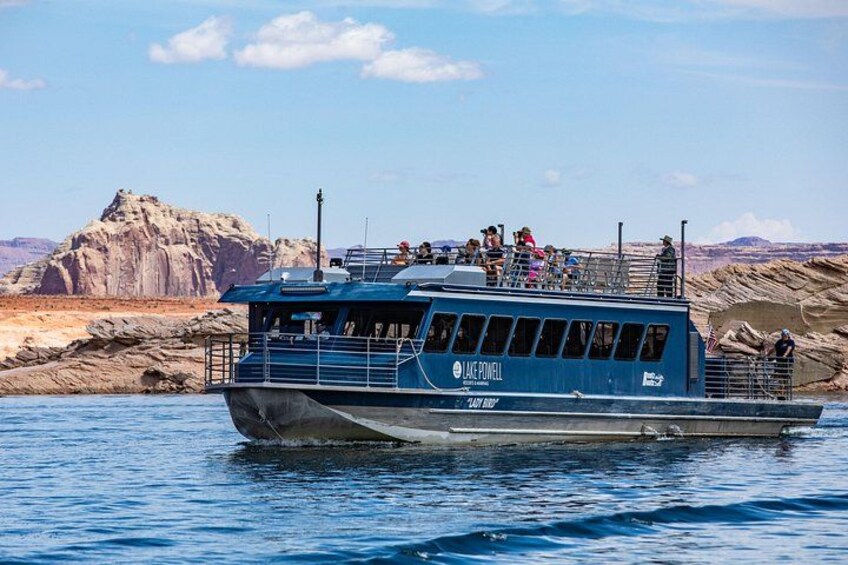 Un bateau bleu, baptisé « Lace Powell », avec des personnes à bord, navigue sur une étendue d’eau, avec un paysage rocheux en arrière-plan. 1