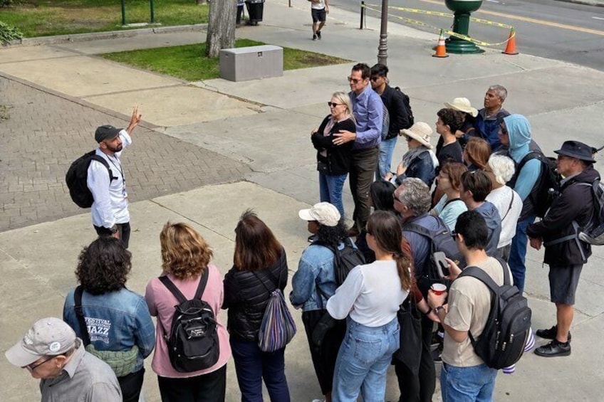 Un groupe de personnes se tient sur un trottoir, certaines avec des sacs à dos, près d’un bac à fleurs vert et d’une rue. 2