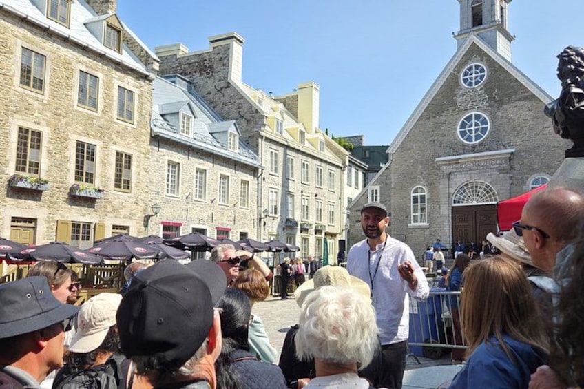 A crowd gathered around a street performer in a historic town square with stone buildings and a church. 1