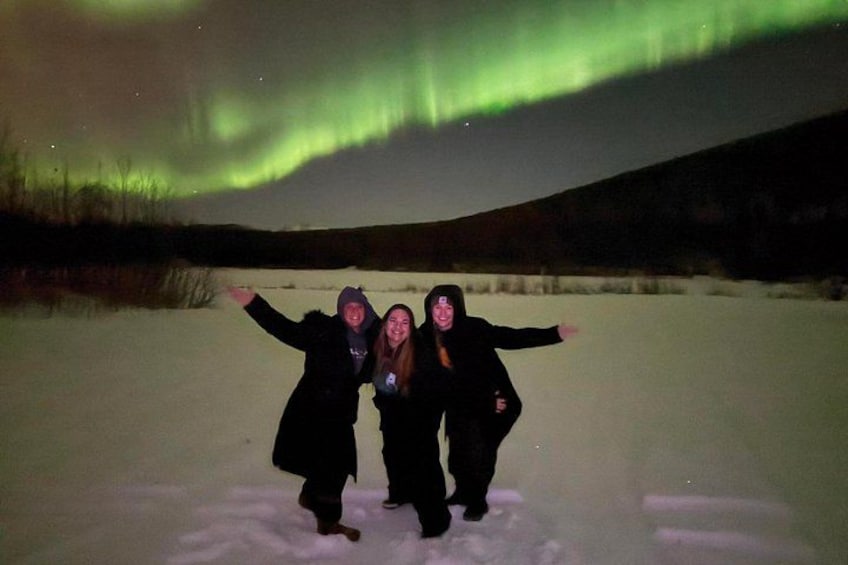 Trois personnes se tiennent dans un champ enneigé, les bras tendus, sous un ciel nocturne illuminé par des aurores boréales vertes. 5