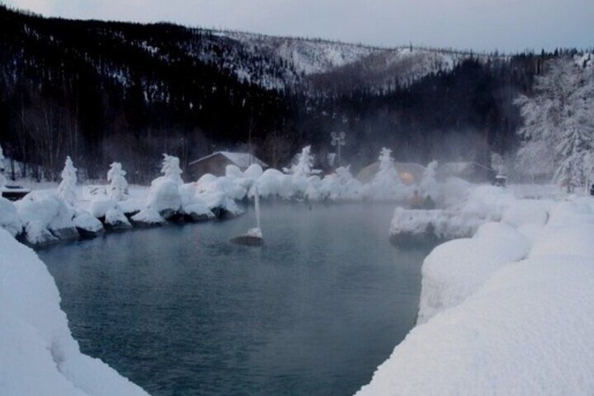 A snow-covered hot spring surrounded by trees and a forested hill. 1