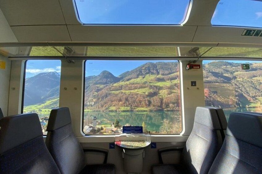 Interior de un tren con asientos y una mesa, vista por la ventana de un paisaje montañoso con un lago. 2