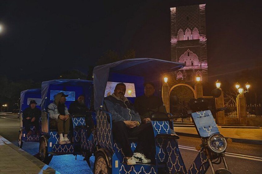 A nighttime scene with a group of people seated in a decorated vehicle, a lit-up tower in the background, and a street with lights. 2