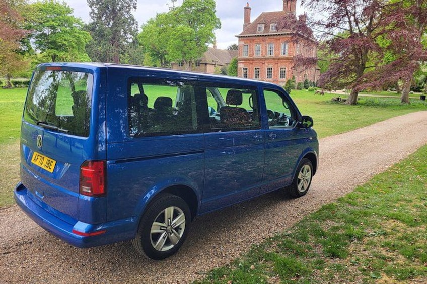 A blue Volkswagen van parked on a gravel path with a red brick building and trees in the background. 2