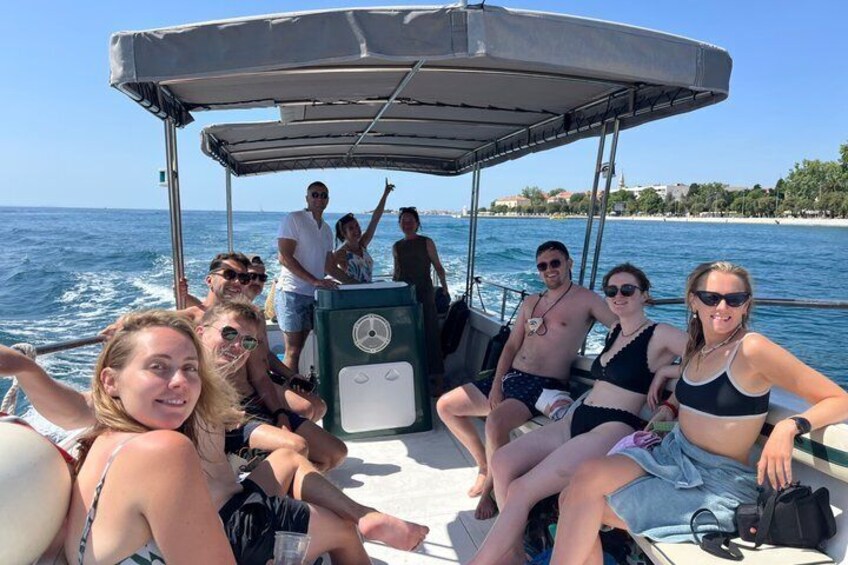 Een groep mensen op een boot met een zonnescherm, water en een strand op de achtergrond. 4
