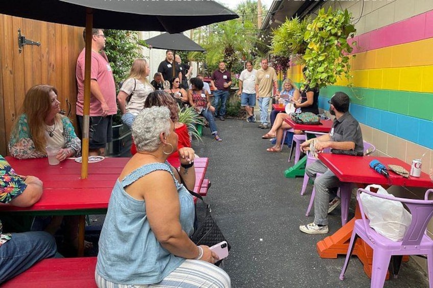 People seated at outdoor tables with colorful chairs and umbrellas, in a casual outdoor setting. 4