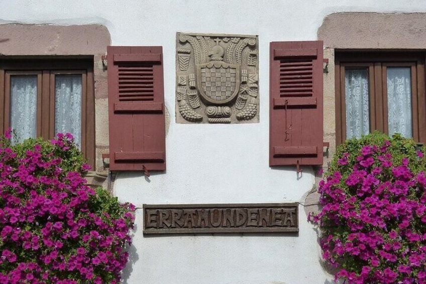 A building facade with a crest emblem, two windows with brown shutters, and a sign reading 'FERRAKUNDENEA'. 1