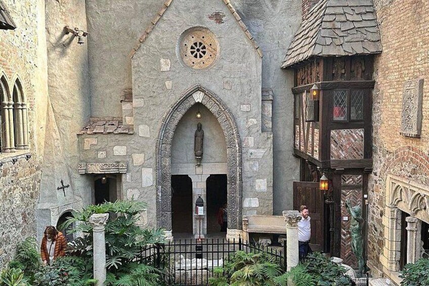 A medieval-style building with a stone facade, a wooden door, and a small garden area with plants and a fence. 5