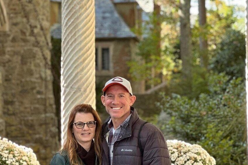 A couple standing in front of a stone building with a large column and flowering bushes. 4