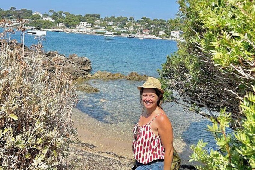 A coastal scene with a person standing on a rocky shore, a clear blue sea, and buildings in the background. 1