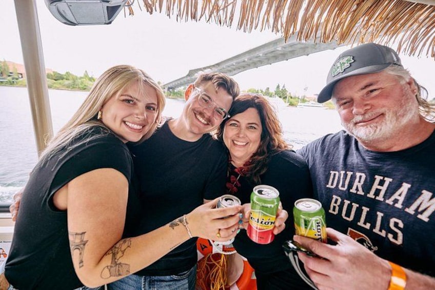 A group of people on a boat, holding drinks, with a river and a bridge in the background. 3